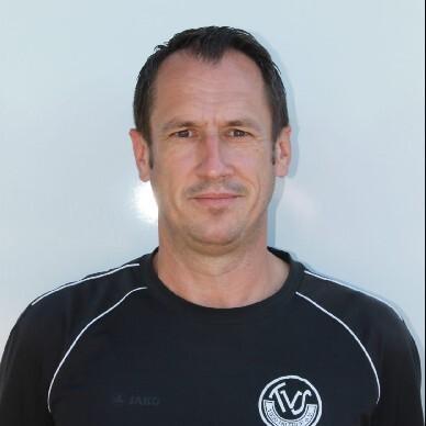 Portr&auml;t eines Mannes in schwarzem Sportshirt mit Vereinslogo, vor einer hellen, neutralen Hintergrundfl&auml;che.