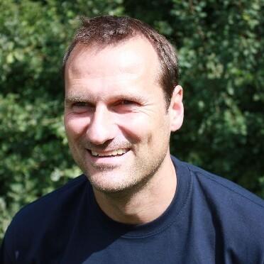 Mann mit kurzem Haar lächelt vor unscharfem, grünem Hintergrund in einem dunkelblauen Shirt.