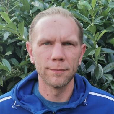 Mann mit kurzen Haaren trägt eine blaue Sportjacke vor einem Hintergrund aus grünen Blättern.