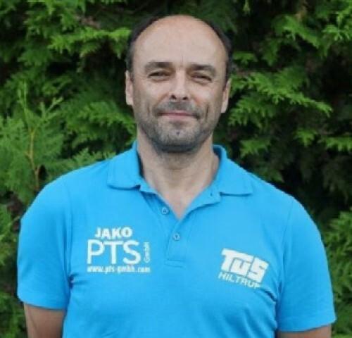 Mann in blauem Poloshirt steht vor grünem Hintergrund aus Pflanzen.