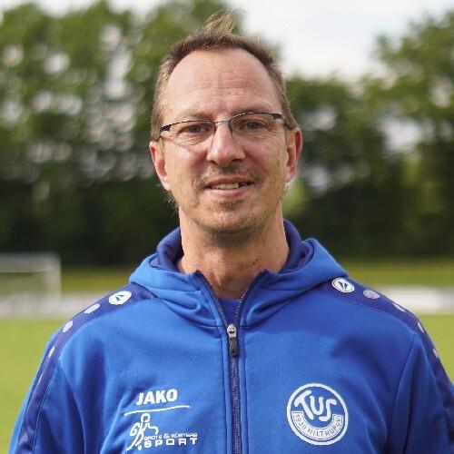 Mann in blauer Trainingsjacke steht lächelnd vor einem Fußballfeld mit unscharfem Hintergrund.
