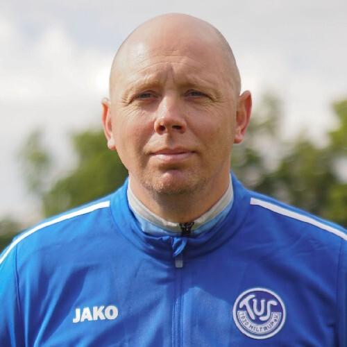 Portr&auml;t eines Mannes in blauer Trainingsjacke mit Logo, vor unscharfer gr&uuml;ner Landschaft.