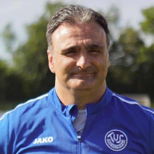 Porträt eines Mannes in blauer Sportjacke mit Vereinslogo, lächelnd vor einem Sportfeld mit Bäumen im Hintergrund.