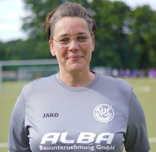 Portrait einer Frau in grauem Sporttrikot, l&auml;chelnd, mit Brille und Stadionhintergrund.