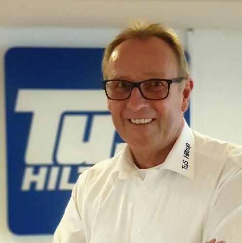 Ein l&auml;chelnder Mann in einem wei&szlig;en Hemd mit Brille steht vor einem blauen Firmenlogo.