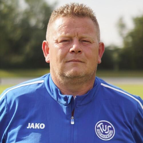 Portr&auml;t eines Trainers in blauer Sportjacke, mit ernstem Ausdruck, vor unscharfem Hintergrund.