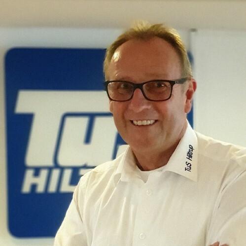 Ein lächelnder Mann in einem weißen Hemd mit Brille steht vor einem blauen Firmenlogo.