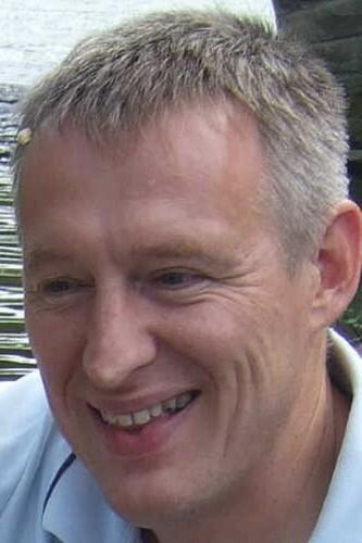 Nahaufnahme eines lächelnden Mannes mit kurzem grauen Haar in natürlicher Umgebung am Wasser.