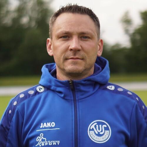 Porträt eines Mannes in blauer Sportjacke mit Vereinslogo, im Freien vor verschwommenem Hintergrund.