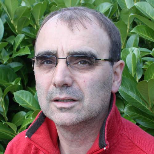 Porträt eines Mannes mit Brille, der in einem roten Pullover vor einem Hintergrund aus grünen Pflanzen steht.