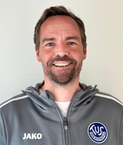 Porträt eines lächelnden Mannes mit kurzem Haar, in grauer Sportjacke mit Logo und Aufschrift „JAKO“.