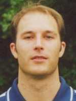 Nahaufnahme eines Mannes mit kurzem Haar, in einem Poloshirt, vor einer grünen, unscharfen Hintergrundlandschaft.