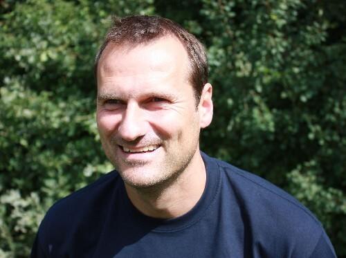 Mann mit kurzem Haar lächelt vor unscharfem, grünem Hintergrund in einem dunkelblauen Shirt.