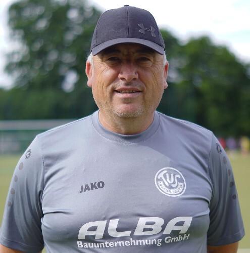 Porträt eines lächelnden Fußballtrainers in grauem Shirt mit Logo und Kappe, Hintergrund mit Bäumen und Sportplatz.