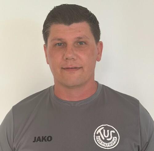 Porträt eines Mannes mit dunklen, kurzen Haaren, der ein graues Sportshirt mit Logo trägt, vor einem weißen Hintergrund.