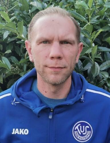 Mann mit kurzen Haaren trägt eine blaue Sportjacke vor einem Hintergrund aus grünen Blättern.