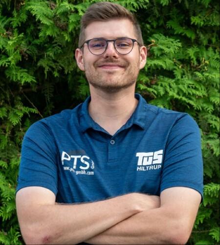 Mann mit Brille und lächelndem Gesicht steht mit verschränkten Armen vor einem grünen Hintergrund aus Pflanzen.