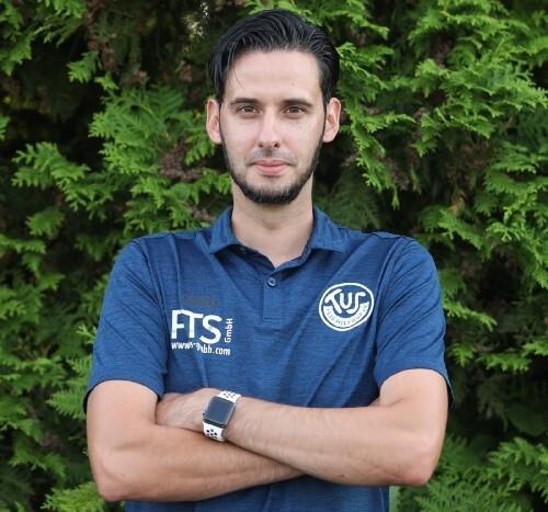 Porträt eines Mannes im blauen Poloshirt mit verschränkten Armen vor einer grünen Heckenlandschaft.