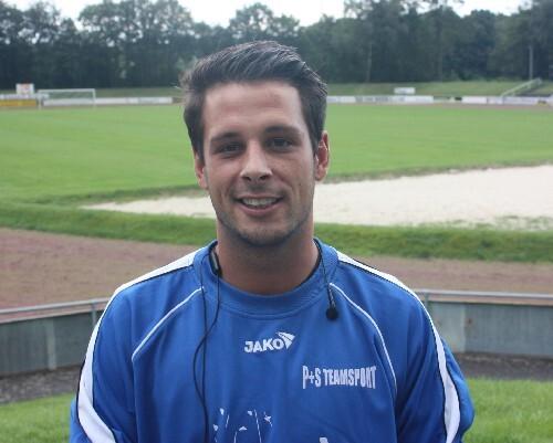 Portrait eines lächelnden Mannes in Sportkleidung, stehend auf einem Fußballplatz mit grünem Rasen im Hintergrund.