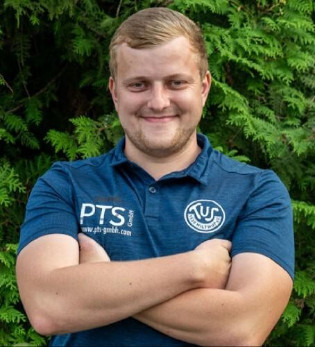 Junger Mann mit kurzem Haar in blauer Sportbekleidung steht lächelnd vor einer grünen Pflanzenwand.