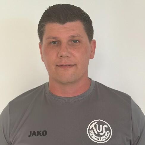 Porträt eines Mannes mit dunklen, kurzen Haaren, der ein graues Sportshirt mit Logo trägt, vor einem weißen Hintergrund.