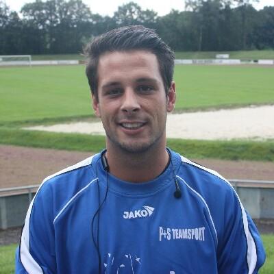 Portrait eines lächelnden Mannes in Sportkleidung, stehend auf einem Fußballplatz mit grünem Rasen im Hintergrund.