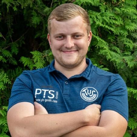 Junger Mann mit kurzem Haar in blauer Sportbekleidung steht lächelnd vor einer grünen Pflanzenwand.
