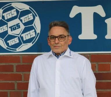 Mann in weißem Hemd steht vor einer roten Backsteinwand mit einem Fußball-Logo und einem Schriftzug.