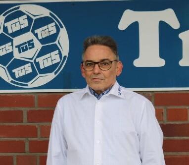 Mann in wei&szlig;em Hemd steht vor einer roten Backsteinwand mit einem Fu&szlig;ball-Logo und einem Schriftzug.
