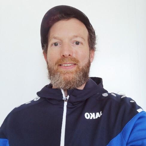 Portrait eines Mannes mit Bart, der einen blauen Sportanzug und eine Kappe trägt, vor einem hellen Hintergrund.