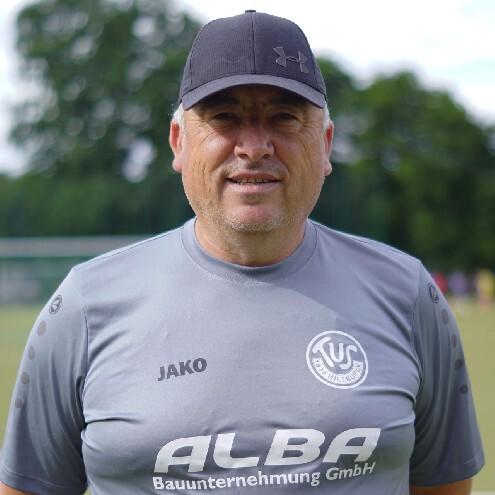 Portr&auml;t eines l&auml;chelnden Fu&szlig;balltrainers in grauem Shirt mit Logo und Kappe, Hintergrund mit B&auml;umen und Sportplatz.