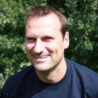 Mann mit kurzem Haar lächelt vor unscharfem, grünem Hintergrund in einem dunkelblauen Shirt.