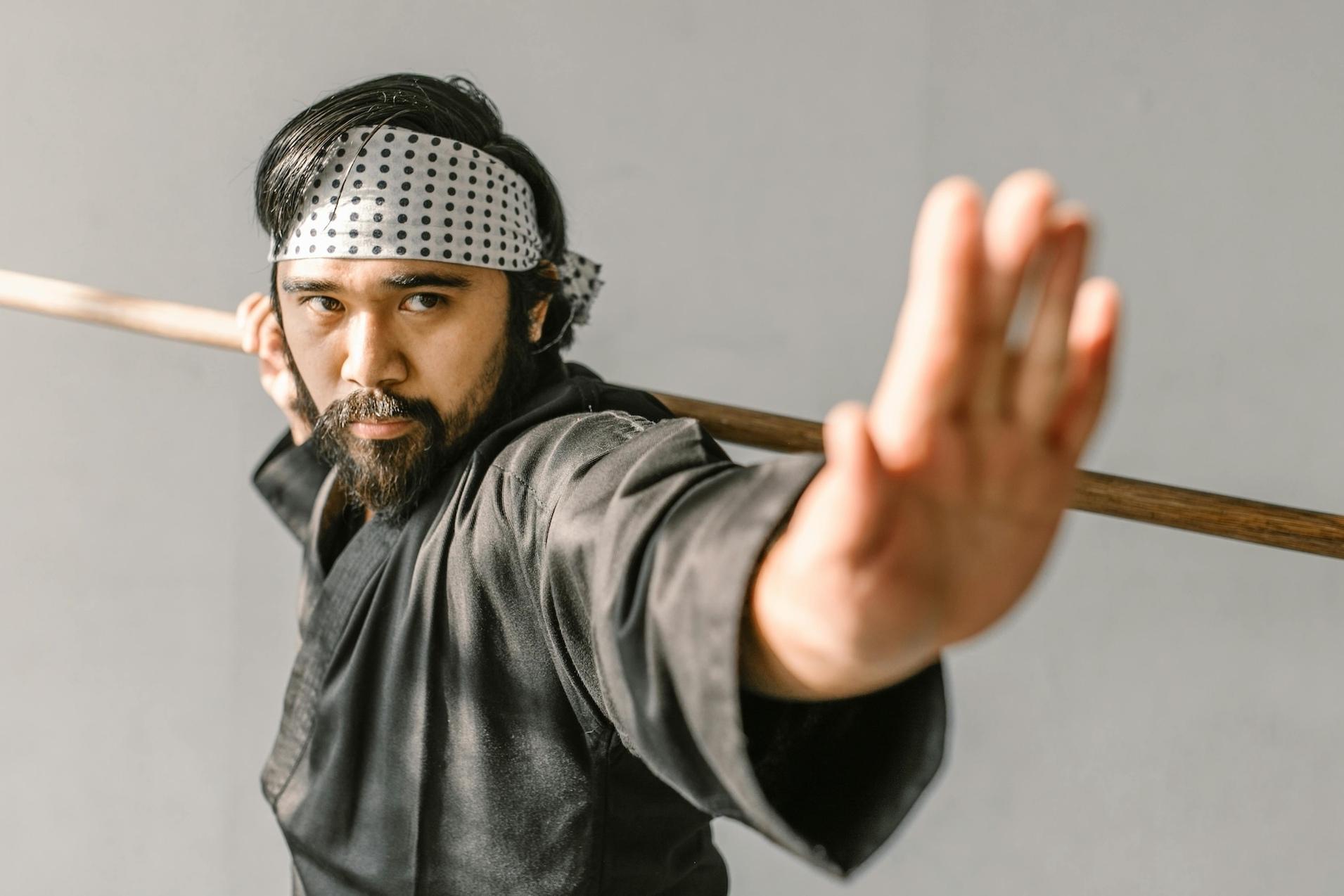 Mann in schwarzem Kimono mit einem Stock, zeigt eine Kampfhaltung in einem neutralen Raum.