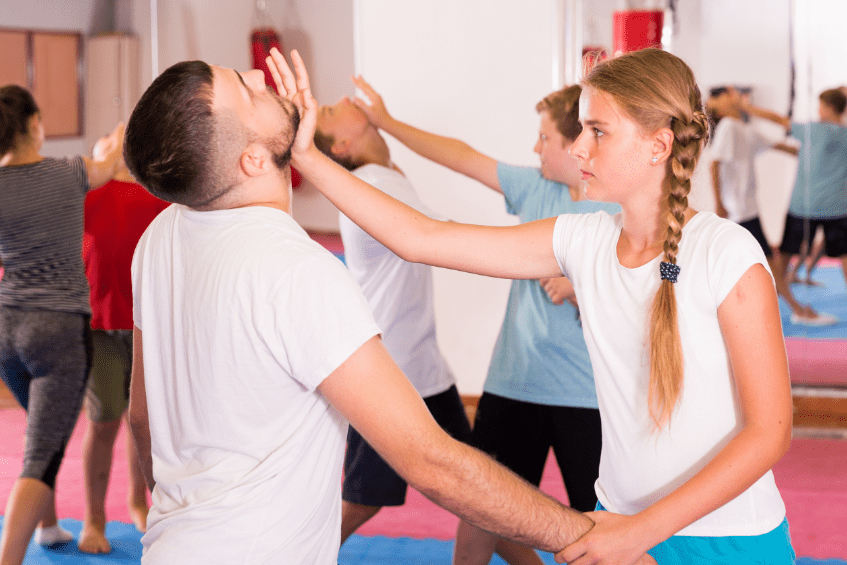 Kurs Rolling Selfdefense Zwei Personen üben Selbstverteidigung in einem Trainingsraum mit Matten und Spiegeln, weitere Teilnehmer im Hintergrund.