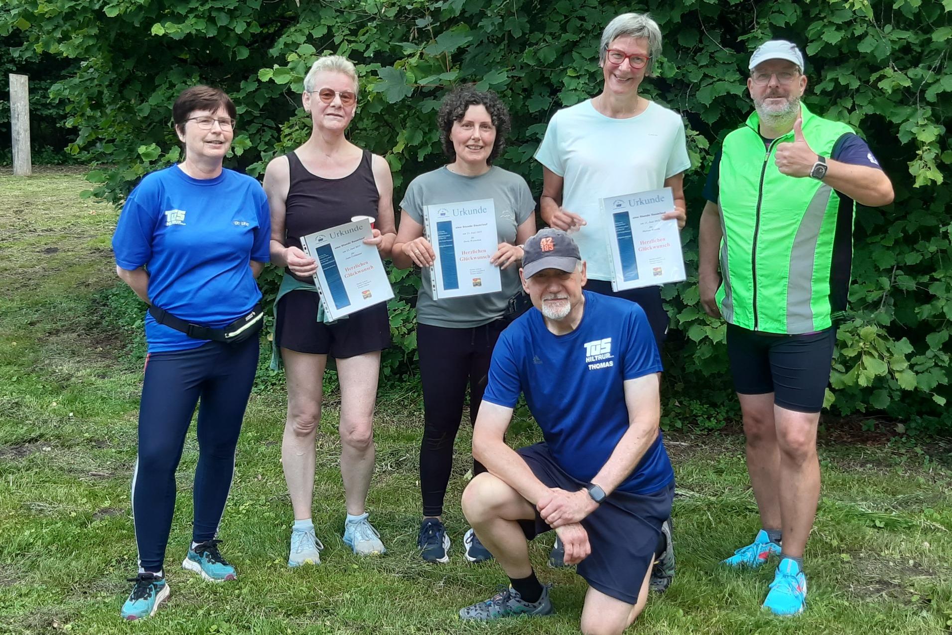 Gruppe von sechs Personen im Freien, die stolz ihre Urkunden in der Hand halten, vor einer grünen Baumkulisse.