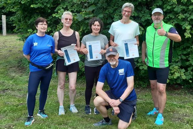 Gruppe von sechs Personen im Freien, die stolz ihre Urkunden in der Hand halten, vor einer grünen Baumkulisse.
