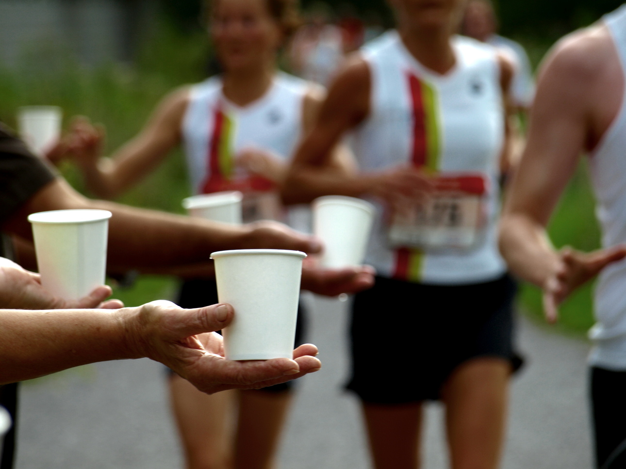 Papiertassen mit Getr&auml;nken werden von mehreren H&auml;nden an laufende Sportler &uuml;berreicht. Hintergrund mit anderen L&auml;ufern.
