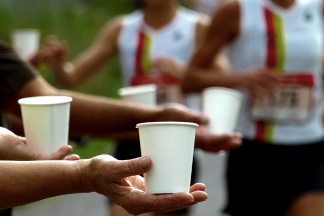 Papiertassen mit Getränken werden von mehreren Händen an laufende Sportler überreicht. Hintergrund mit anderen Läufern.