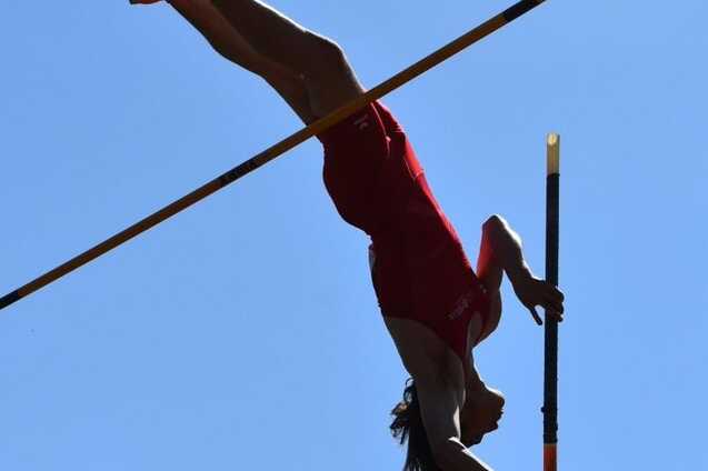 Sportlerin beim Stabhochsprung, kopfüber über eine Stange vor blauem Himmel, in rotem Outfit und orangefarbenen Schuhen.