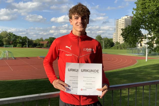 Sportler in roter Jacke h&auml;lt Urkunden auf einem Sportplatz mit Rasen und Laufbahn im Hintergrund.