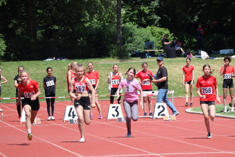 Sechs junge Athletinnen bei einem Wettlauf auf einer roten Tartanbahn, Zuschauer im Hintergrund.