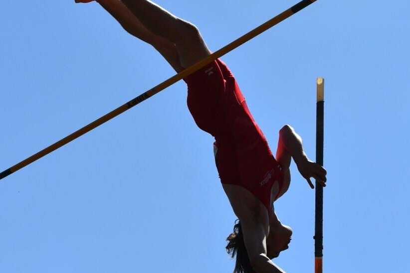 Sportlerin beim Stabhochsprung, kopfüber über eine Stange vor blauem Himmel, in rotem Outfit und orangefarbenen Schuhen.