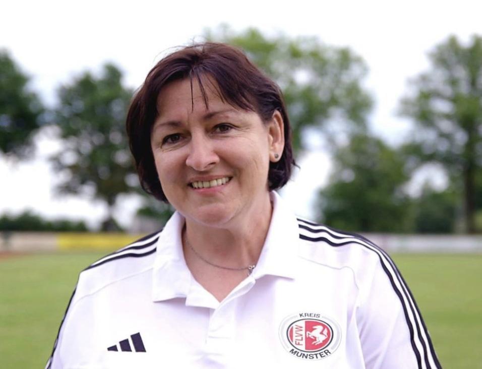 Portr&auml;t einer l&auml;chelnden Frau in einem wei&szlig;en Shirt mit Vereinslogo, stehend auf einem Sportfeld mit B&auml;umen im Hintergrund.