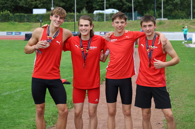 Vier jubelnde junge Athleten in roten Trikots mit Medaillen stehen auf einem Sportplatz und posieren für ein Foto.