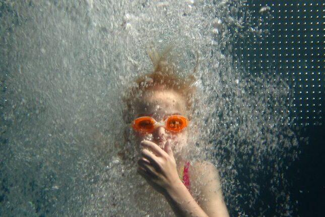 Mädchen mit orangefarbener Schwimmbrille taucht unter Wasser, während Blasen aufsteigen.