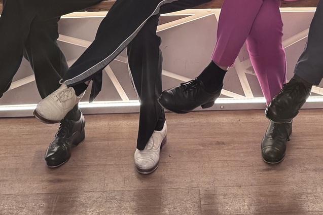 Reihe von F&uuml;&szlig;en in verschiedenen Schuhen, die &uuml;ber einer Holzoberfl&auml;che h&auml;ngen, teils mit bunten Socken.