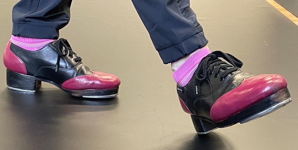 Nahaufnahme eines Fußes in einem schwarzen und pinken Stepptanzschuh auf einem dunklen Boden.