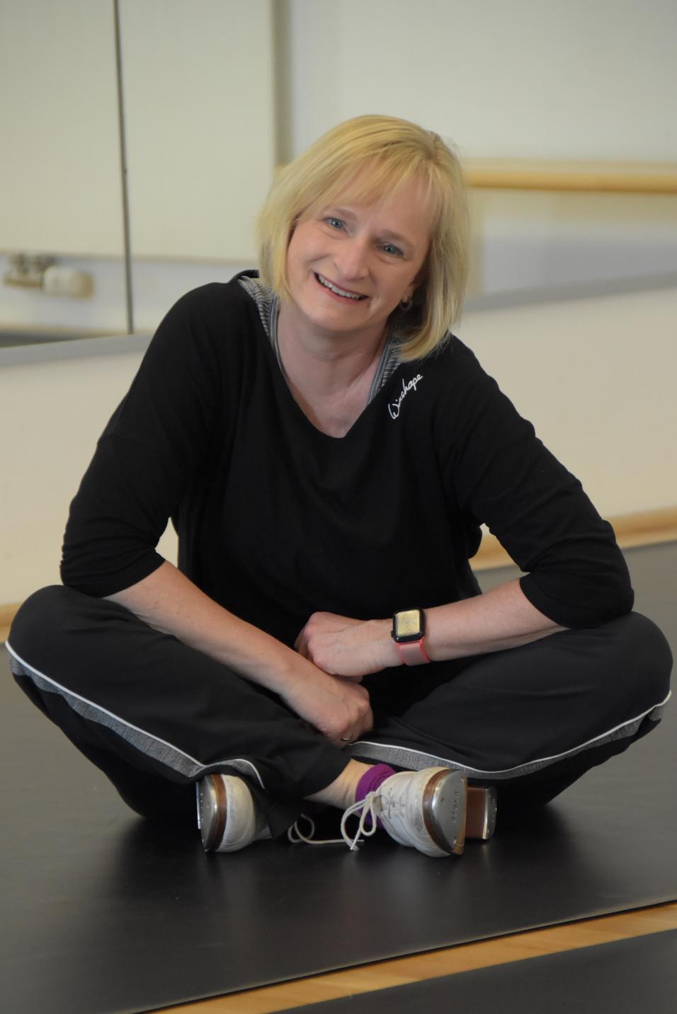 Frau in Sportkleidung sitzt mit gekreuzten Beinen auf einem schwarzen Boden und lächelt in einem Fitnessraum.