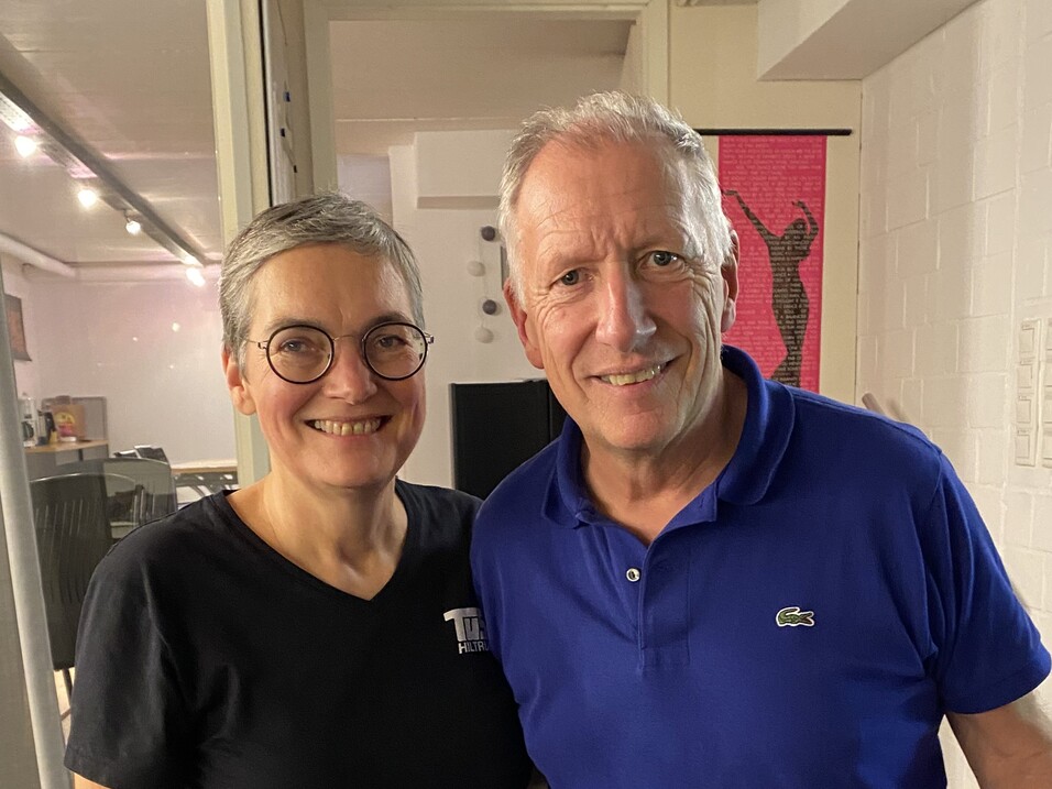 Paar lächelt in einem Raum, die Frau trägt ein schwarzes T-Shirt und grüne Hosen, der Mann ein blaues Polohemd.