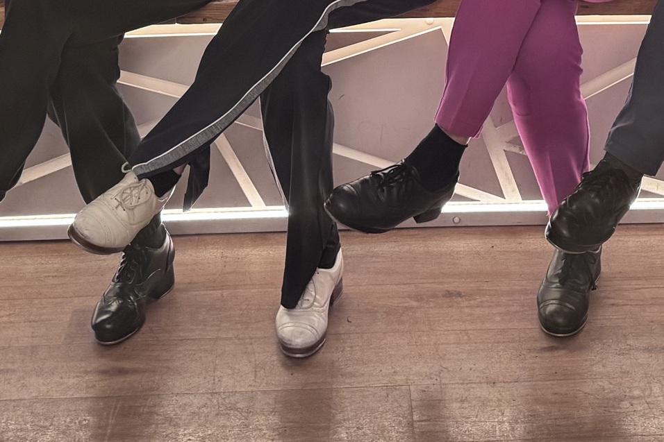 Reihe von F&uuml;&szlig;en in verschiedenen Schuhen, die &uuml;ber einer Holzoberfl&auml;che h&auml;ngen, teils mit bunten Socken.